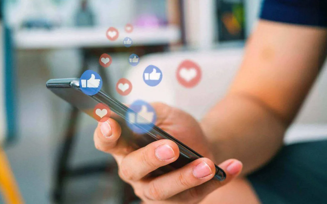 Pessoa usando o celular com símbolos de curtidas e reações nas redes sociais.